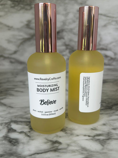 Two yellow frosted glass bottles of Revelry Crafts Moisturizing Body Mist with rose gold caps, standing on a white and gray marble surface. One bottle shows the front label reading 'Believe' and the other shows the side label.