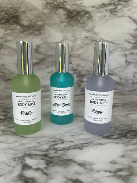 Three bottles of body mist arranged vertically on a white and gray marble surface. The bottles are frosted in gray, teal, and sage green, with silver caps and white labels reading 'BODY MIST' and 'Rogue', 'After Dark', and 'Riddle' respectively.