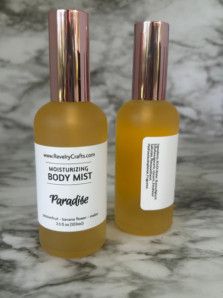 Two gold frosted glass bottles of Revelry Crafts Paradise Moisturizing Body Mist, showing the front and back labels, topped with metallic rose gold caps, set on a gray and white marble surface.
