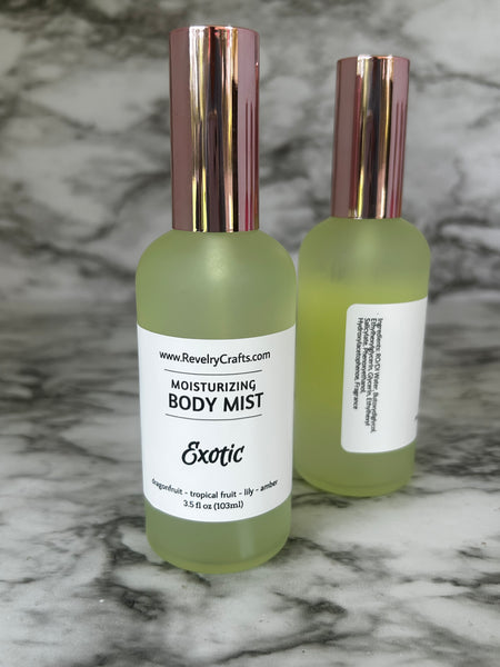 Two yellow frosted glass bottles of Exotic Moisturizing Body Mist with rose gold caps, displayed on a gray and white marble surface.
