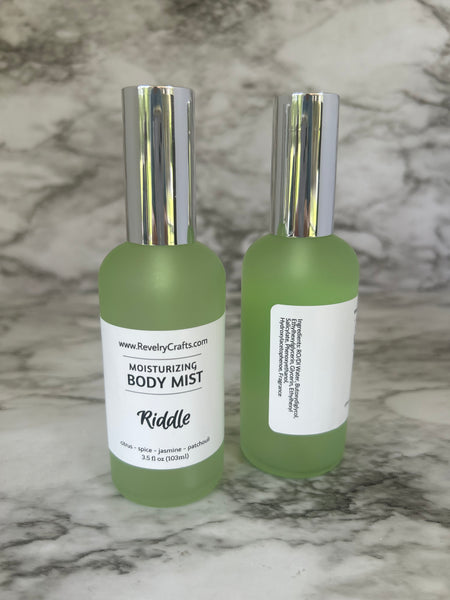 Two frosted light green bottles of Riddle Moisturizing Body Mist with silver caps, displayed on a gray and white marble surface.