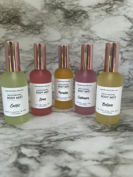 Five frosted glass body mist bottles with rose gold caps arranged vertically on a gray and white marble surface. The bottles contain liquids in different muted colors and have white labels with black text reading 'Moisturizing Body Mist' with the names of each scent beneath it.
