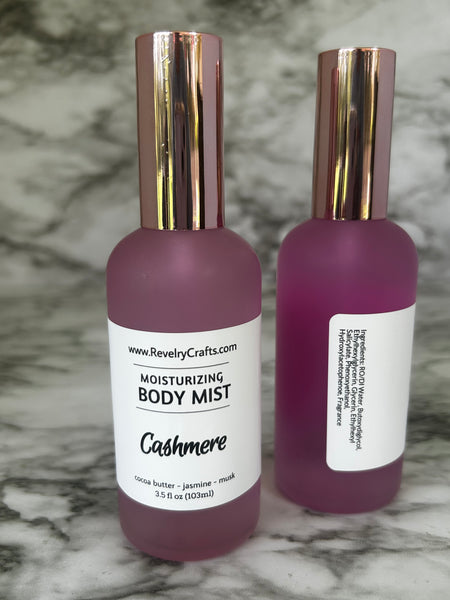 Two pink frosted glass body mist bottles with metallic rose gold caps, showing the front and back labels for Revelry Crafts Cashmere Moisturizing Body Mist on a gray marble surface.